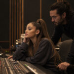 Woman seated at a large audio mixing console looking at the computer screen displaying a DAW as a man leans over to help in a dimly lit recording studio