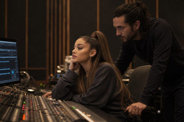 Woman seated at a large audio mixing console looking at the computer screen displaying a DAW as a man leans over to help in a dimly lit recording studio