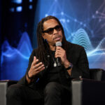 Man with dreadlocks in black attire sits on stage in a chair, speaking into a microphone and gesturing, with a blue digital backdrop behind him.