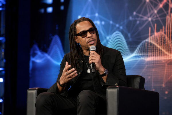 Man with dreadlocks in black attire sits on stage in a chair, speaking into a microphone and gesturing, with a blue digital backdrop behind him.