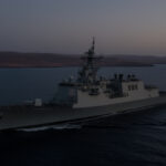 Gray military destroyer at sea during sunset coastline visible on the horizon