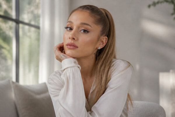 Portrait of a woman with long blonde hair in a white blouse, resting her chin on her hand in a bright living space, looking at the camera.