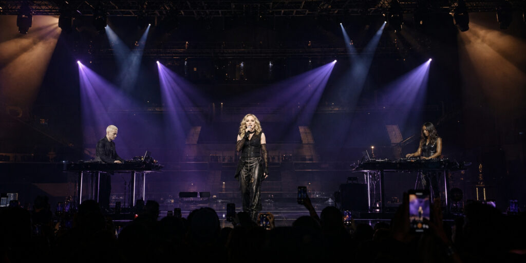 Female singer on a dark stage under purple spotlights, flanked by two DJs at electronic consoles; audience holds up phones in silhouette.