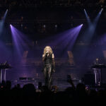 Female singer on a dark stage under purple spotlights, flanked by two DJs at electronic consoles; audience holds up phones in silhouette.