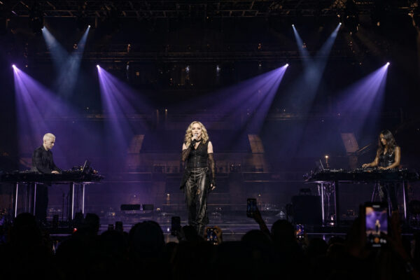 Female singer on a dark stage under purple spotlights, flanked by two DJs at electronic consoles; audience holds up phones in silhouette.