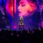 Pop artist center stage singing in a black outfit, two musicians at sides, large neon-lit backdrop with a close-up on screen behind the singer, crowd filming with phones below.