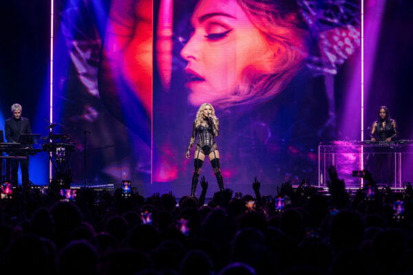 Pop artist center stage singing in a black outfit, two musicians at sides, large neon-lit backdrop with a close-up on screen behind the singer, crowd filming with phones below.