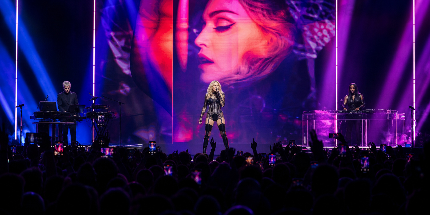 Pop artist center stage singing in a black outfit, two musicians at sides, large neon-lit backdrop with a close-up on screen behind the singer, crowd filming with phones below.